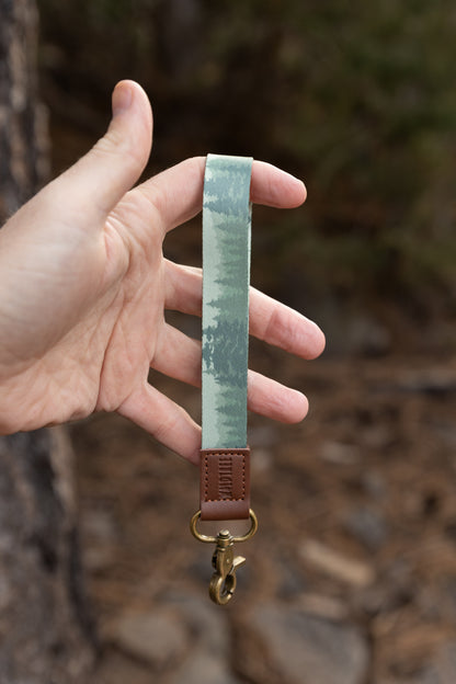 A close-up shot of a person's hand holding a wristlet keychain vertically. The strap features a "foggy forest" pattern with light green and grey pine tree silhouettes. It is finished with a brown leather accent embossed with "WILDTREE" and a brass-colored metal lobster clasp.