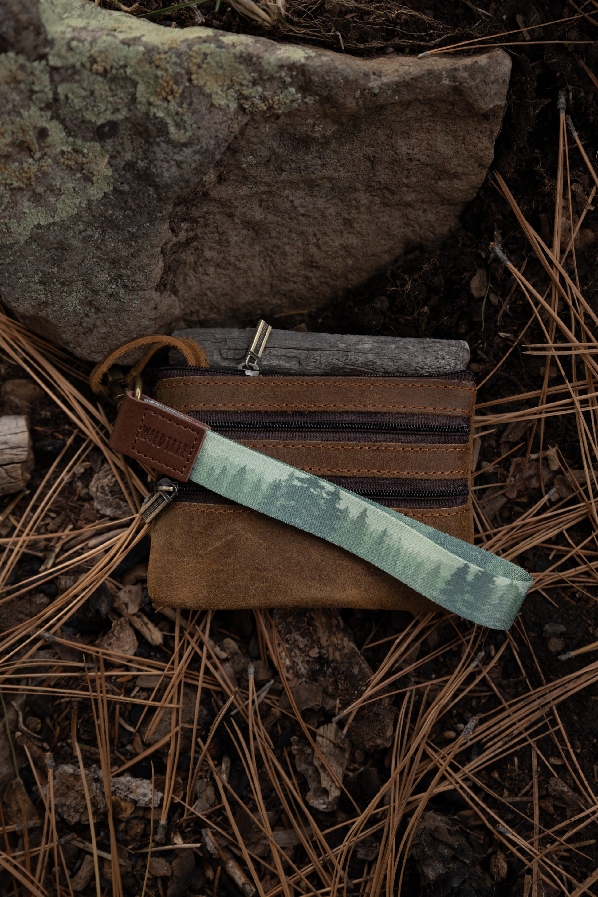A foggy forest wristlet keychain is attached to a small, dark brown leather zippered pouch or wallet. The items are resting on a forest floor covered in pine needles and dirt, positioned against a mossy rock.