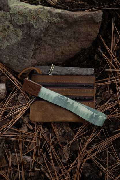 A foggy forest wristlet keychain is attached to a small, dark brown leather zippered pouch or wallet. The items are resting on a forest floor covered in pine needles and dirt, positioned against a mossy rock.