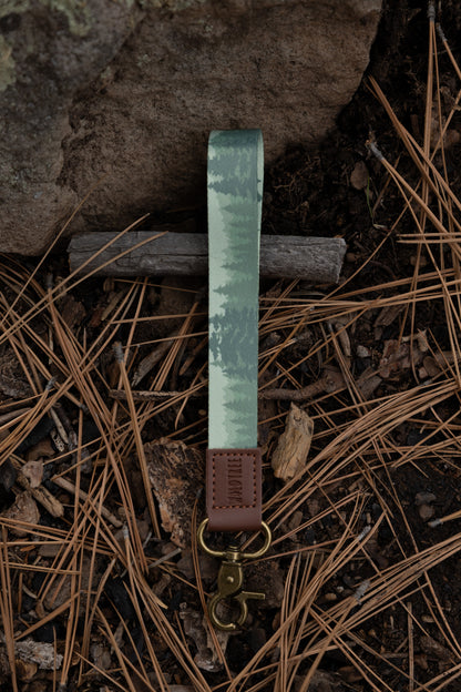 The foggy forest wristlet keychain is laid flat on a bed of dry pine needles and forest soil next to a large rock. The strap points upward, showcasing the gradient of evergreen tree silhouettes against a misty background.