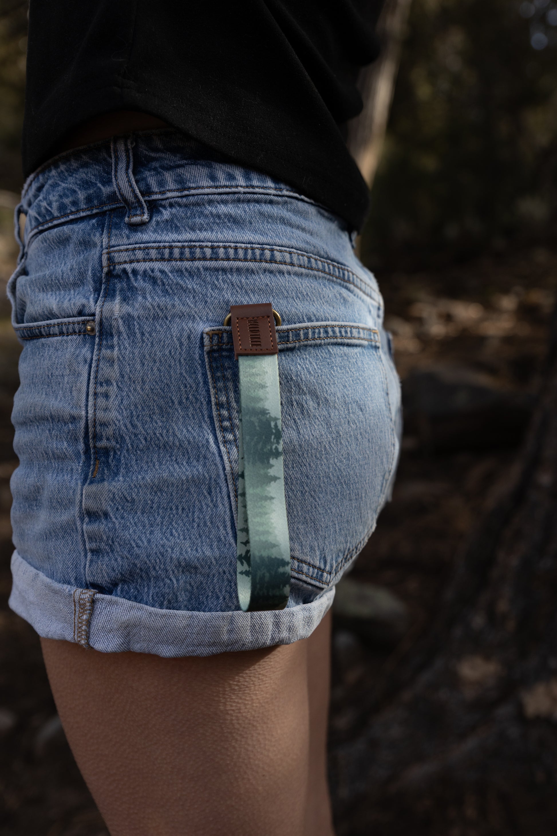 A close-up view from behind a person wearing blue jeans, with a forest-patterned wristlet keychain hanging from their back pocket. The brown leather tab of the keychain sits at the edge of the pocket, and the strap showcases a hazy, atmospheric pine tree design.