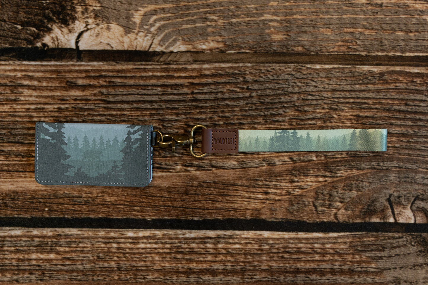 The Foggy Forest wristlet keychain shown on a rustic wooden background, clipped to a small, matching fabric SD card holder that features the same misty evergreen forest landscape design.