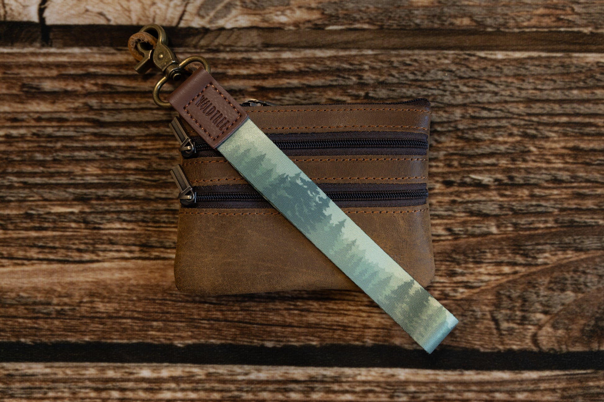 A lifestyle shot on a wood background showing the forest-themed wristlet keychain attached to the top corner of a small, brown leather multi-zippered coin pouch or wallet.