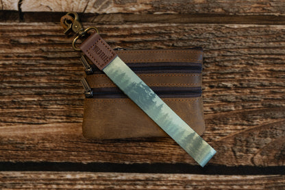 A lifestyle shot on a wood background showing the forest-themed wristlet keychain attached to the top corner of a small, brown leather multi-zippered coin pouch or wallet.