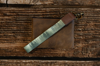 A close-up view of the Foggy Forest wristlet keychain lying diagonally across a brown leather pouch, highlighting the detail of the pine tree print and the durable stitching on the leather end.