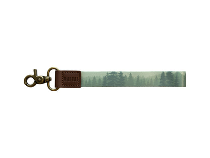 A product photo against a plain white background showing a wristlet keychain with a misty green forest print on a polyester strap, a brown leather connector stamped with "WILDTREE," and an antique bronze swivel clasp.