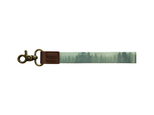A product photo against a plain white background showing a wristlet keychain with a misty green forest print on a polyester strap, a brown leather connector stamped with "WILDTREE," and an antique bronze swivel clasp.