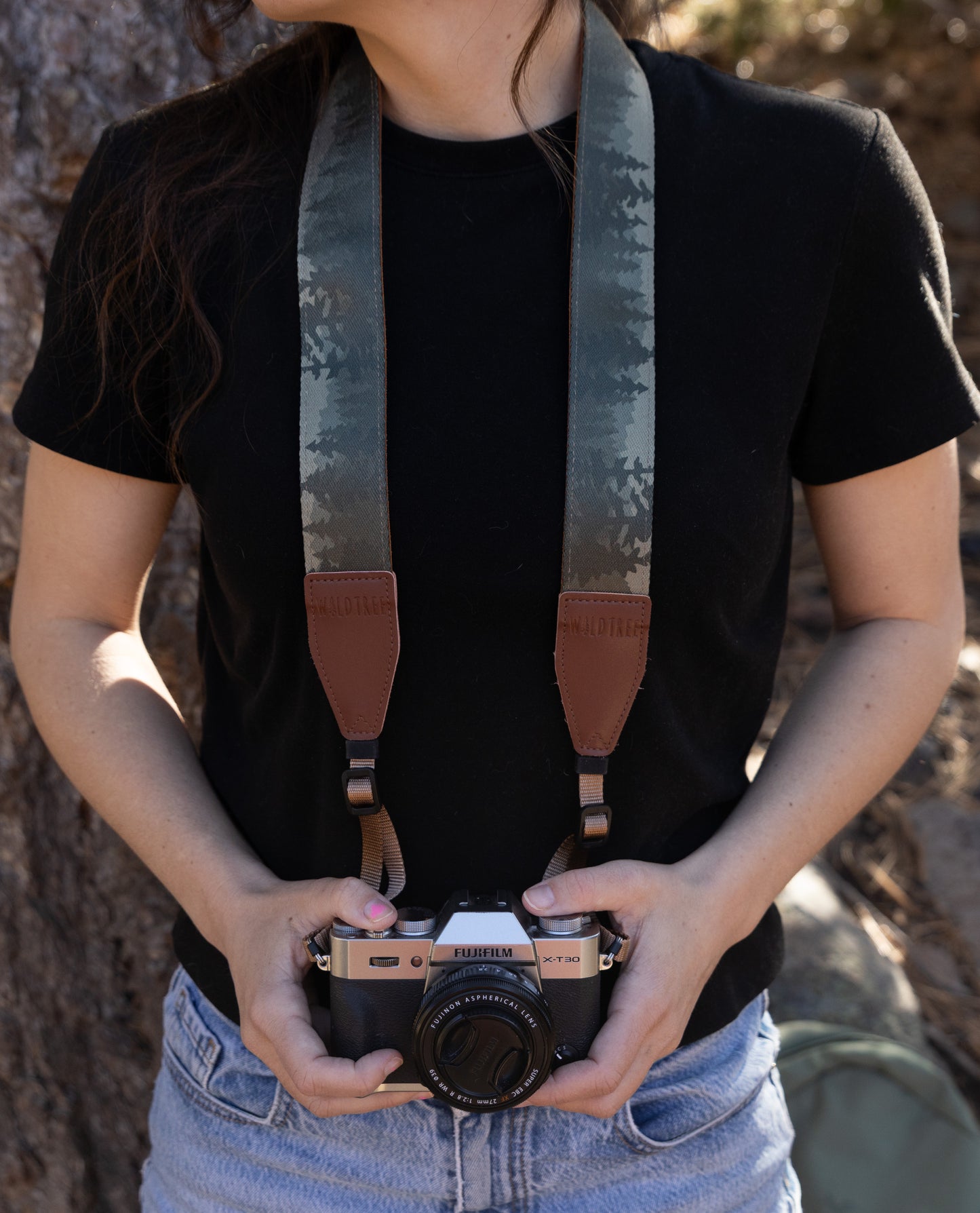 A front-facing, medium shot of a person wearing a black t-shirt and blue denim jeans, holding a silver and black Fujifilm X-T30 camera at waist level. The camera is attached to a wide, fabric camera strap featuring a "foggy forest" print with silhouettes of pine trees in shades of grey and muted green. The strap has brown leather end-tabs embossed with the "WILDTREE" logo.