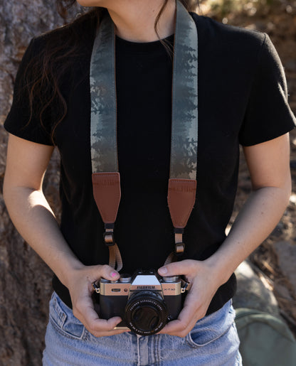 A front-facing, medium shot of a person wearing a black t-shirt and blue denim jeans, holding a silver and black Fujifilm X-T30 camera at waist level. The camera is attached to a wide, fabric camera strap featuring a "foggy forest" print with silhouettes of pine trees in shades of grey and muted green. The strap has brown leather end-tabs embossed with the "WILDTREE" logo.