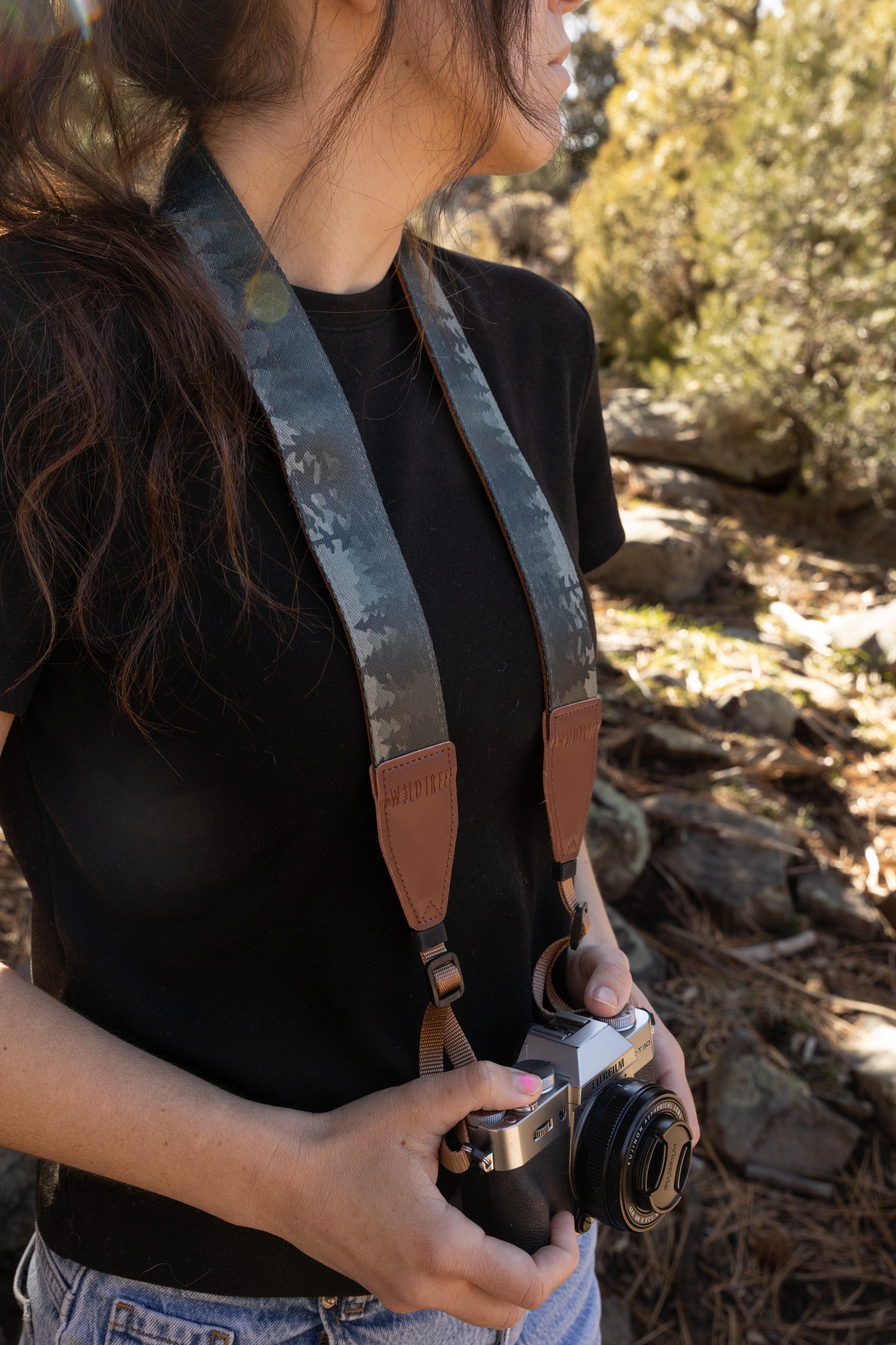 A side-profile view of a person outdoors wearing a black shirt and a camera strap with a dark, misty pine forest pattern. The person is holding a vintage-style mirrorless camera with both hands. The background is a soft-focus forest floor with dry pine needles and rocks, illuminated by warm, dappled sunlight.