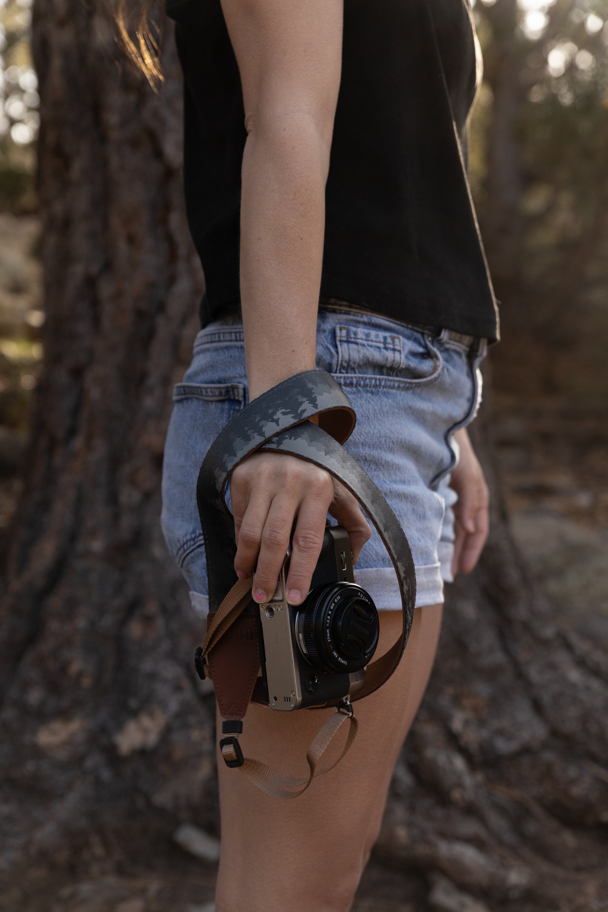A close-up shot of a person’s hand and hip, demonstrating the camera strap wrapped around their wrist for security. The person is wearing denim shorts and holding the silver Fujifilm camera by their side. The "foggy forest" design of the strap is clearly visible, showing the gradient of dark trees against a lighter grey background.