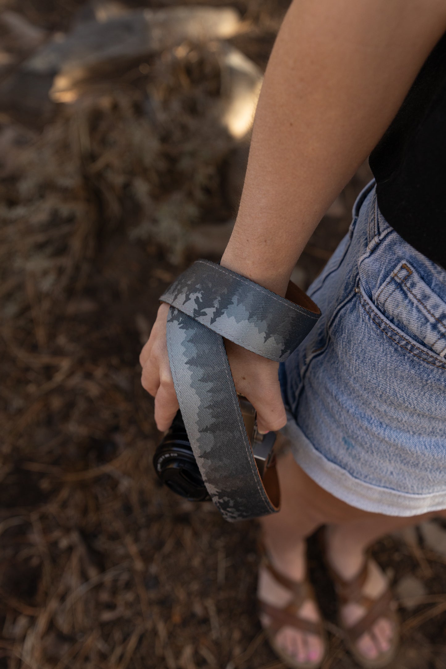 A top-down perspective showing a camera strap wrapped tightly around a person's wrist like a wrist strap. The intricate pine tree silhouette pattern on the strap is the focal point, transitioning from deep charcoal to light misty grey. The person's hand holds the camera, and their brown leather sandals and the forest floor are visible in the blurred background.