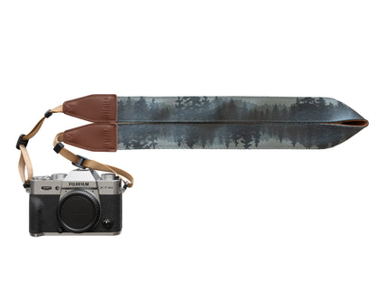 A high-resolution studio shot on a plain white background featuring a silver Fujifilm X-T30 camera attached to the Foggy Forest strap. The strap is laid out horizontally to showcase the full, continuous print of misty evergreen trees and mountains in shades of teal, grey, and charcoal.