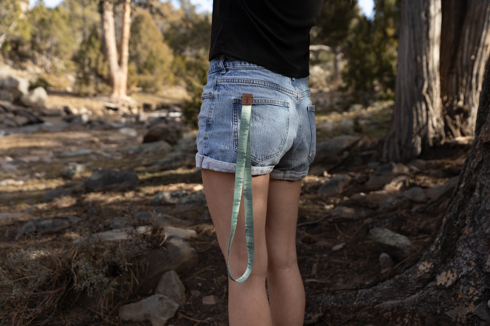 A woman stands in a rugged, sunlit outdoor forest setting, seen from behind. She is wearing denim shorts and a black top. A long, green "Foggy Forest" patterned lanyard is tucked into her back pocket, hanging down against her leg.