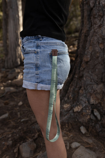 A side-profile view of a person in denim shorts and a black shirt standing in a forest. The green lanyard, featuring a pine tree silhouette pattern, is secured to a belt loop or pocket with a brown leather accent, dangling toward the knee.