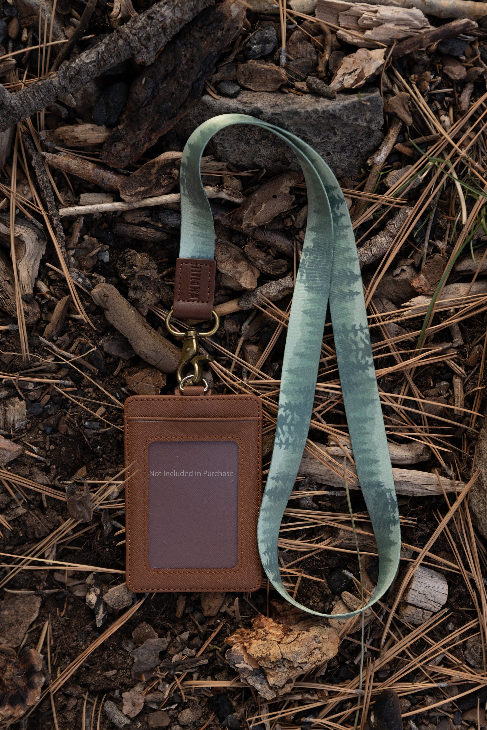 A top-down view of the Foggy Forest lanyard laying on a forest floor covered in pine needles and twigs. Attached to the antique brass swivel clasp is a brown leather-style ID card holder. The card holder's window contains a small note that reads "Not Included in Purchase".