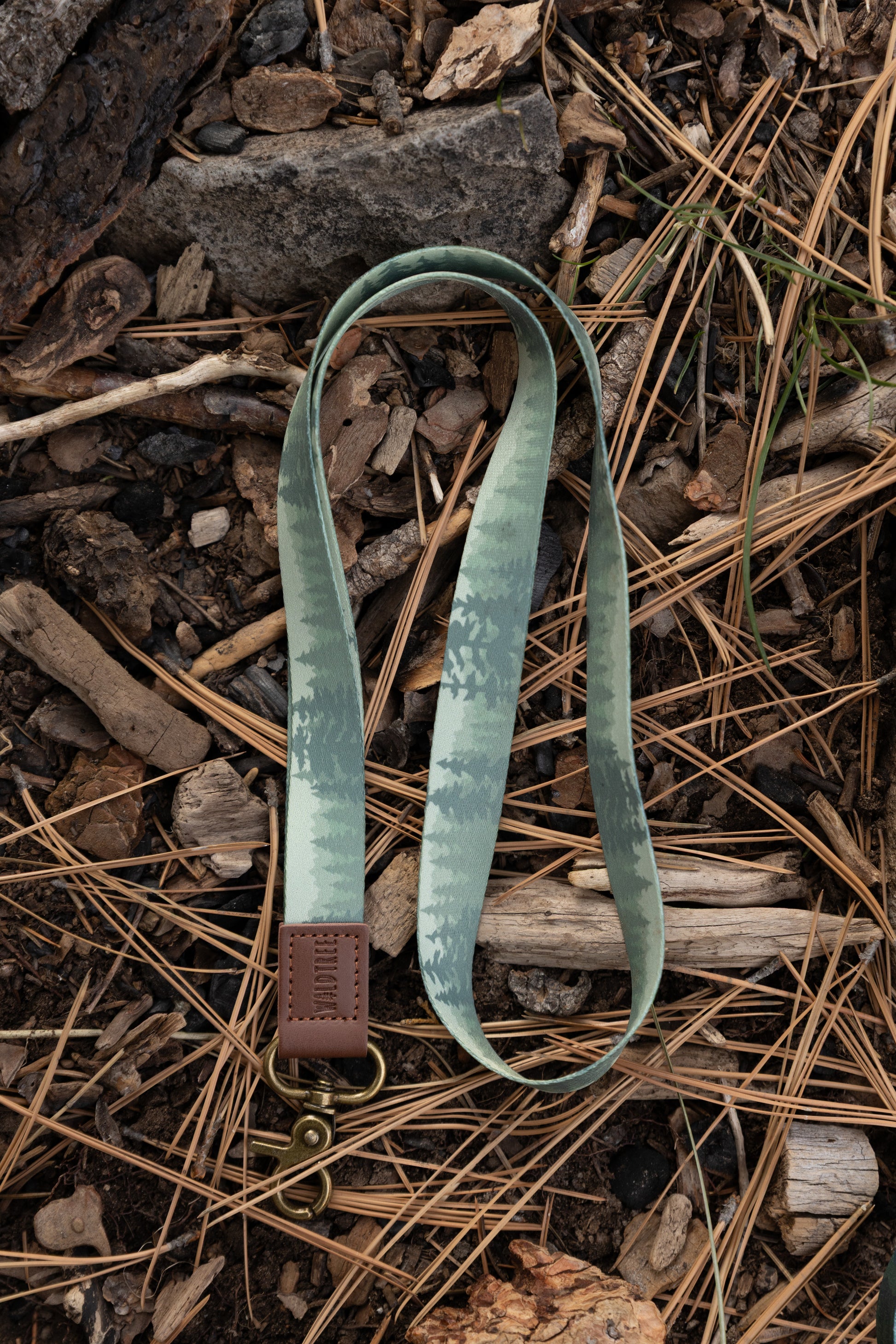 A top-down close-up of the Foggy Forest lanyard resting on a bed of pine needles, twigs, and small rocks. The lanyard features a muted green forest print, a brown leather "WILDTREE" embossed patch, and an antique brass-style swivel lobster clasp.