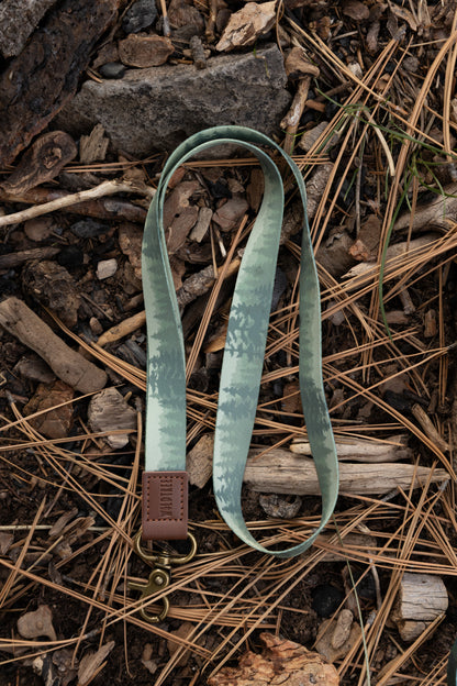 A top-down close-up of the Foggy Forest lanyard resting on a bed of pine needles, twigs, and small rocks. The lanyard features a muted green forest print, a brown leather "WILDTREE" embossed patch, and an antique brass-style swivel lobster clasp.