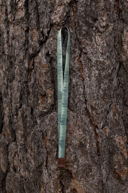 The green forest-patterned lanyard hangs vertically from a small nail embedded in the thick, textured bark of a large pine tree. The lanyard is shown in full length, highlighting the gradient tree design and the leather attachment at the bottom.