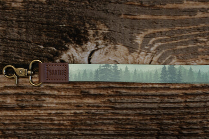 A macro horizontal shot focusing on the branding and hardware of the lanyard. It highlights the "WILDTREE" brand name embossed on a brown leather square, the antique brass-colored metal clasp, and the detailed pine tree silhouettes on the fabric.