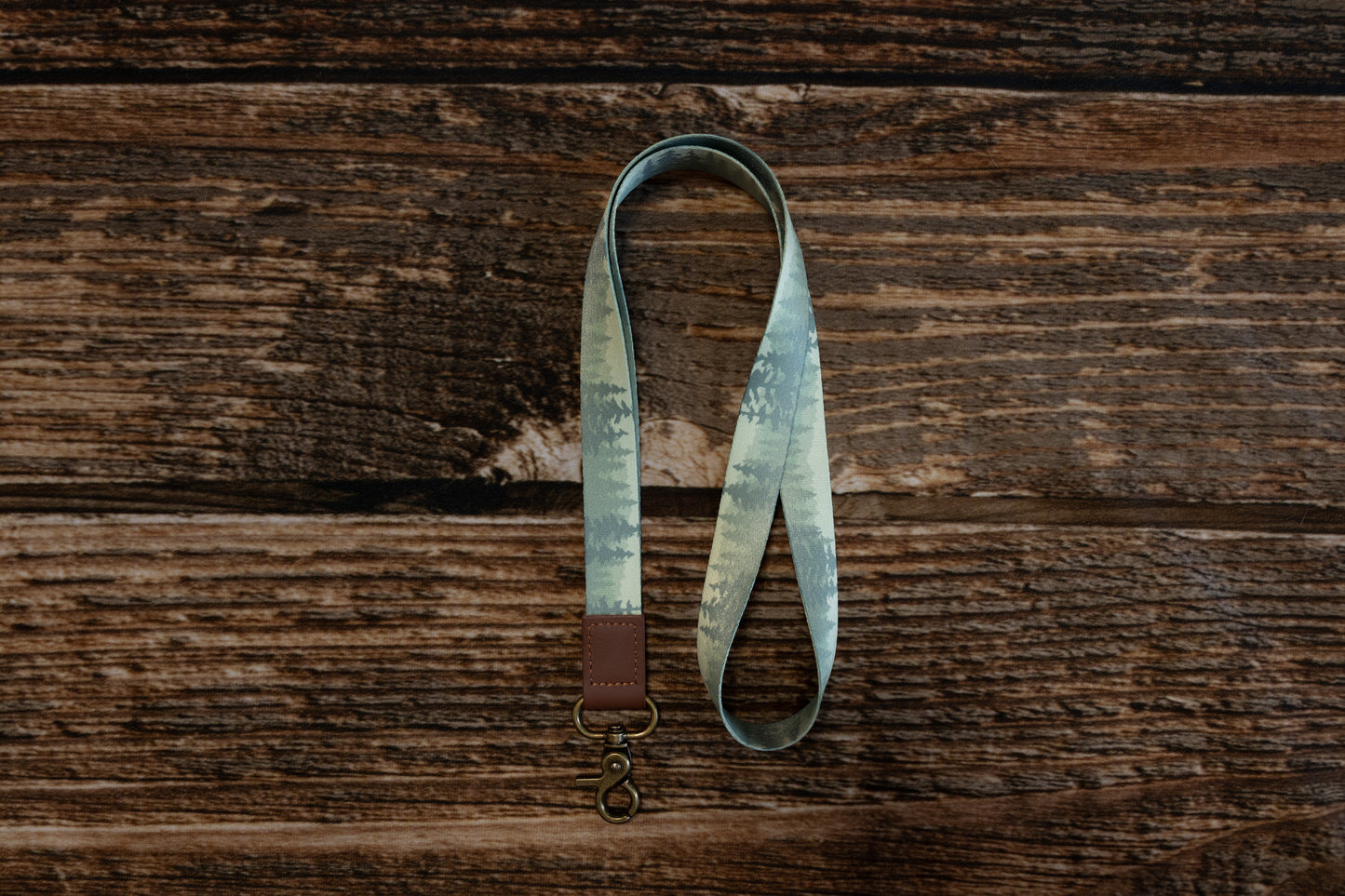 A top-down, vertical view of the forest-patterned lanyard looped on a dark wood background. The pattern showcases layers of green and grey evergreen tree silhouettes. The bottom of the lanyard is finished with a brown leather tab and a brass lobster clasp.