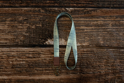 A top-down, vertical view of the forest-patterned lanyard looped on a dark wood background. The pattern showcases layers of green and grey evergreen tree silhouettes. The bottom of the lanyard is finished with a brown leather tab and a brass lobster clasp.