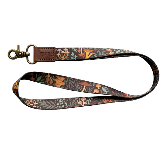 The Forest Foliage Neck Lanyard is displayed against a plain white background. The dark lanyard features a detailed and colorful pattern of various mushrooms in orange, brown, and white, interspersed with green and blue leaves and berries.