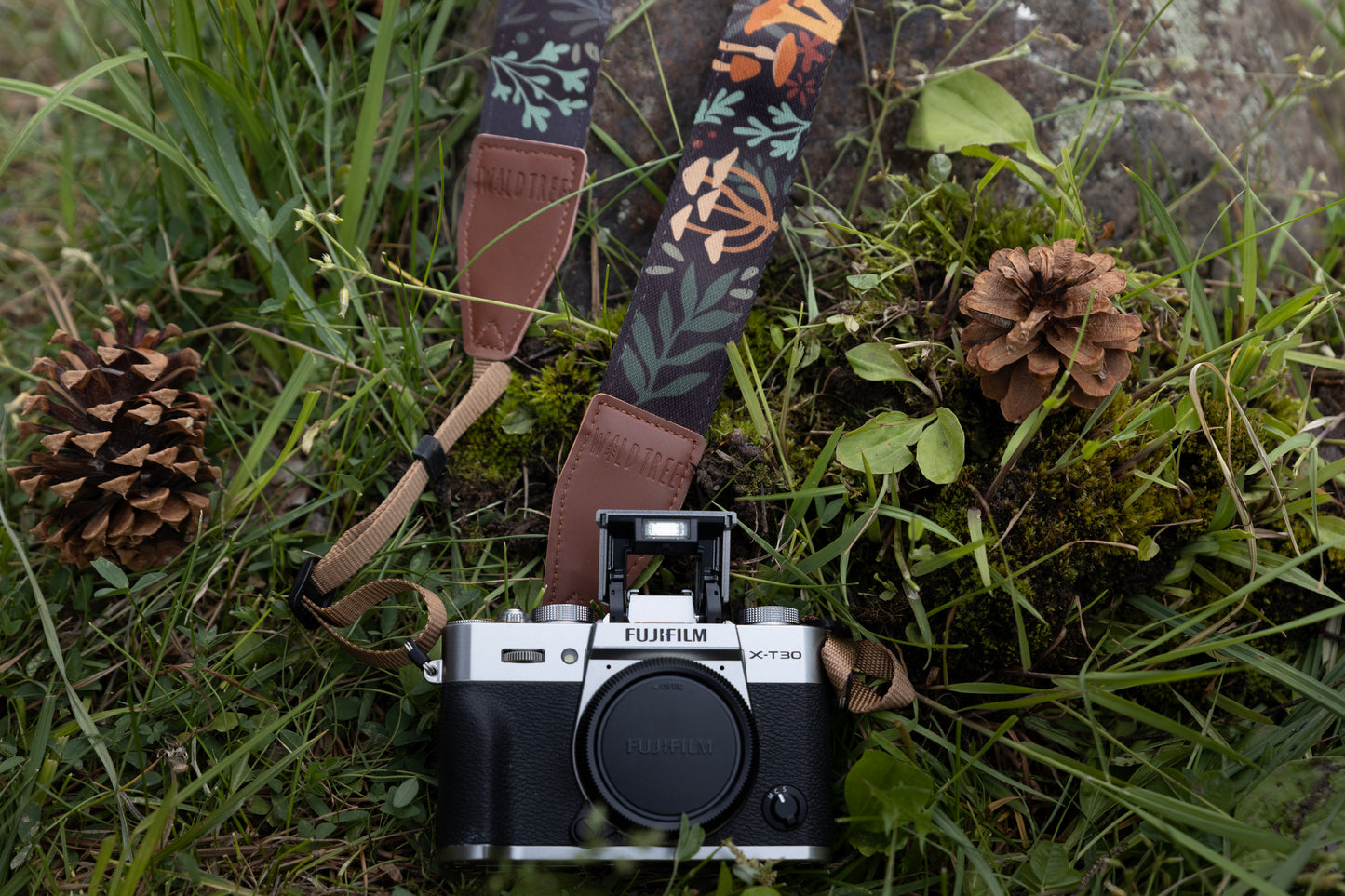 wildtree's forest foliage designed camera strap laying on rocks and moss connected to fujifilm camera