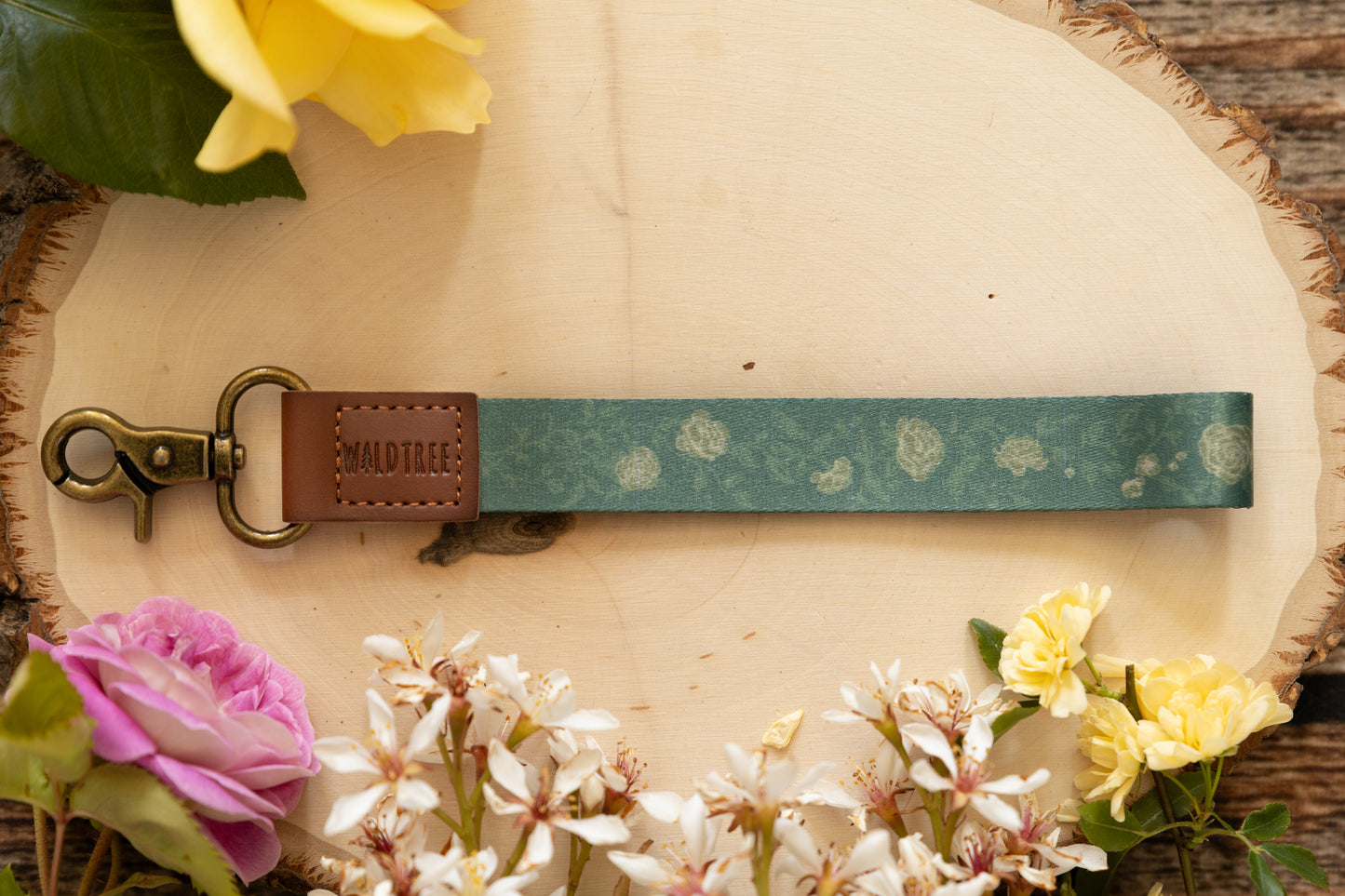 Floral Green Wristlet keychain printed with green vines and yellow flowers laying on wood surrounded by decorative flowers