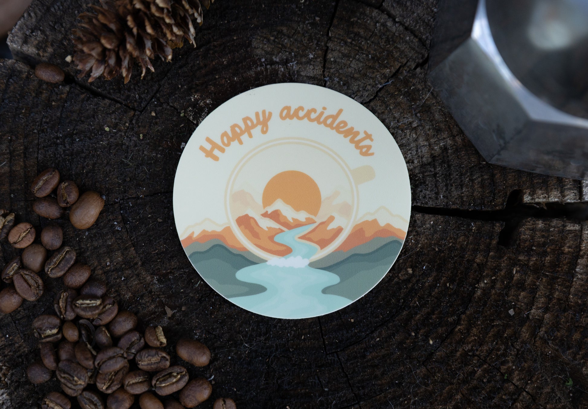A lifestyle photograph taken outdoors, showing the "Happy Accidents" campfire mug decal applied to the rough bark texture of a tree stump. The sticker features an explosive burst of ochre and yellow. Scattered coffee beans and pinecones sit at the base of the wood surface.
