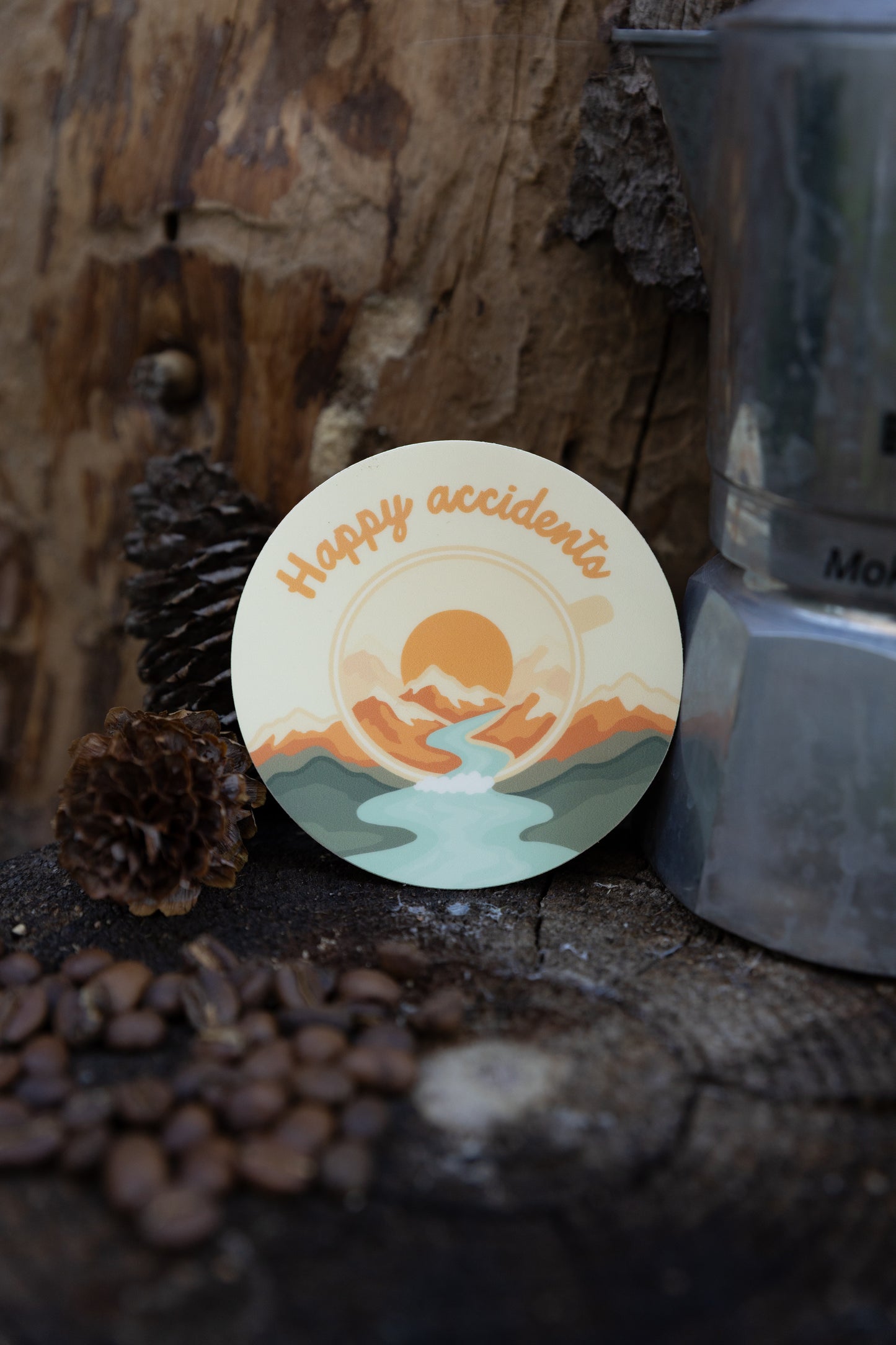 A product showcase photograph of the "Happy Accidents" abstract campfire mug sticker applied to a matte, muted sage green stainless steel travel tumbler. The tumbler is set against a blurred rustic wood background with coffee beans and pinecones at the base.