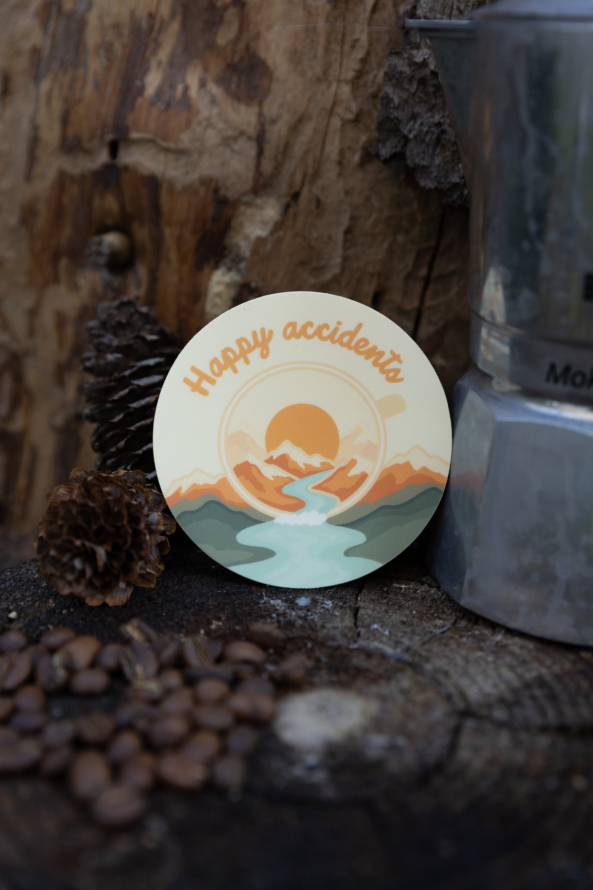A product showcase photograph of the "Happy Accidents" abstract campfire mug sticker applied to a matte, muted sage green stainless steel travel tumbler. The tumbler is set against a blurred rustic wood background with coffee beans and pinecones at the base.