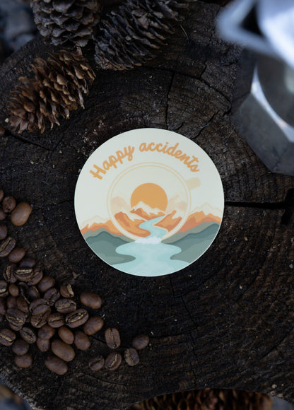 A high-angle flat lay photograph of a die-cut vinyl sticker resting on a rustic wood tree stump. The sticker features an olive green campfire mug with an abstract orange and mustard yellow "splatter" effect exploding from the top, with the text "HAPPY ACCIDENTS" arched below. The scene is framed by dried pinecones and scattered dark roasted coffee beans.