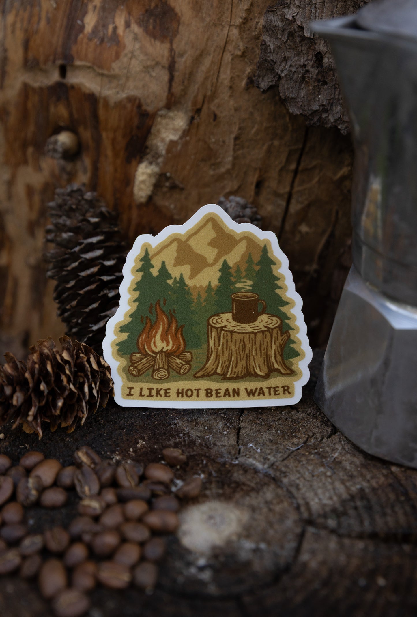 A lifestyle photo of a die-cut vinyl sticker featuring a campfire scene and the text "I LIKE HOT BEAN WATER." The sticker is propped on a dark wood stump next to coffee beans, pinecones, and a silver Moka pot.