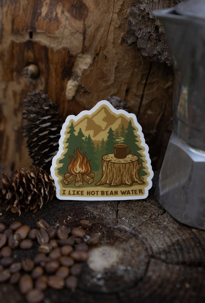 A lifestyle photo of a die-cut vinyl sticker featuring a campfire scene and the text "I LIKE HOT BEAN WATER." The sticker is propped on a dark wood stump next to coffee beans, pinecones, and a silver Moka pot.