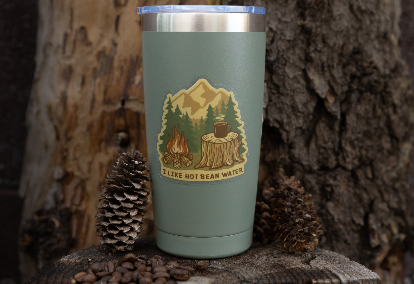 The "Hot Bean Water" sarcastic camping sticker applied to a sage green travel tumbler, shown outdoors on a rustic tree slice with coffee beans and pinecones at the base.