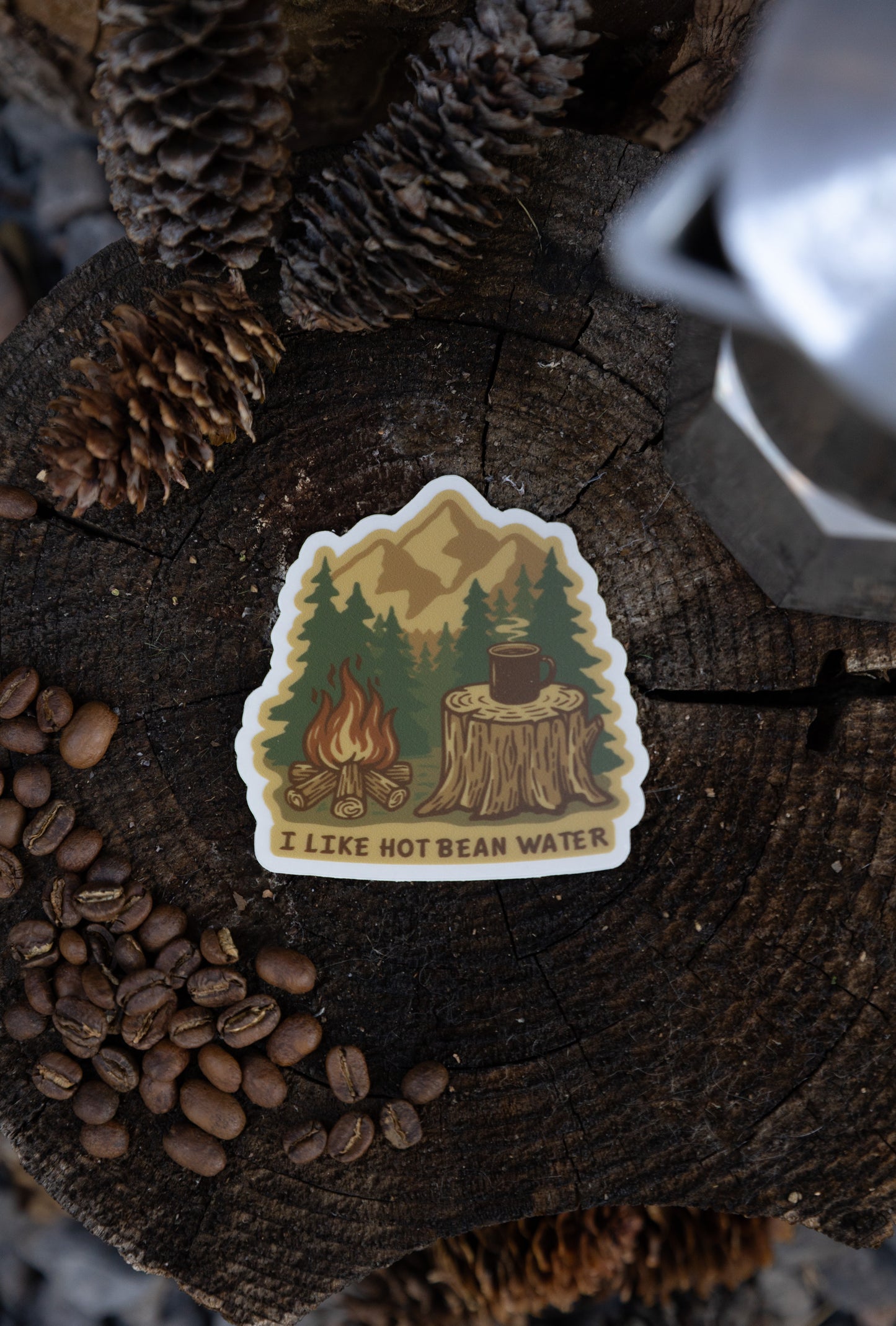 An overhead flat lay of the "I Like Hot Bean Water" sticker resting on a weathered tree stump, surrounded by pinecones, coffee beans, and a silver metal coffee maker.