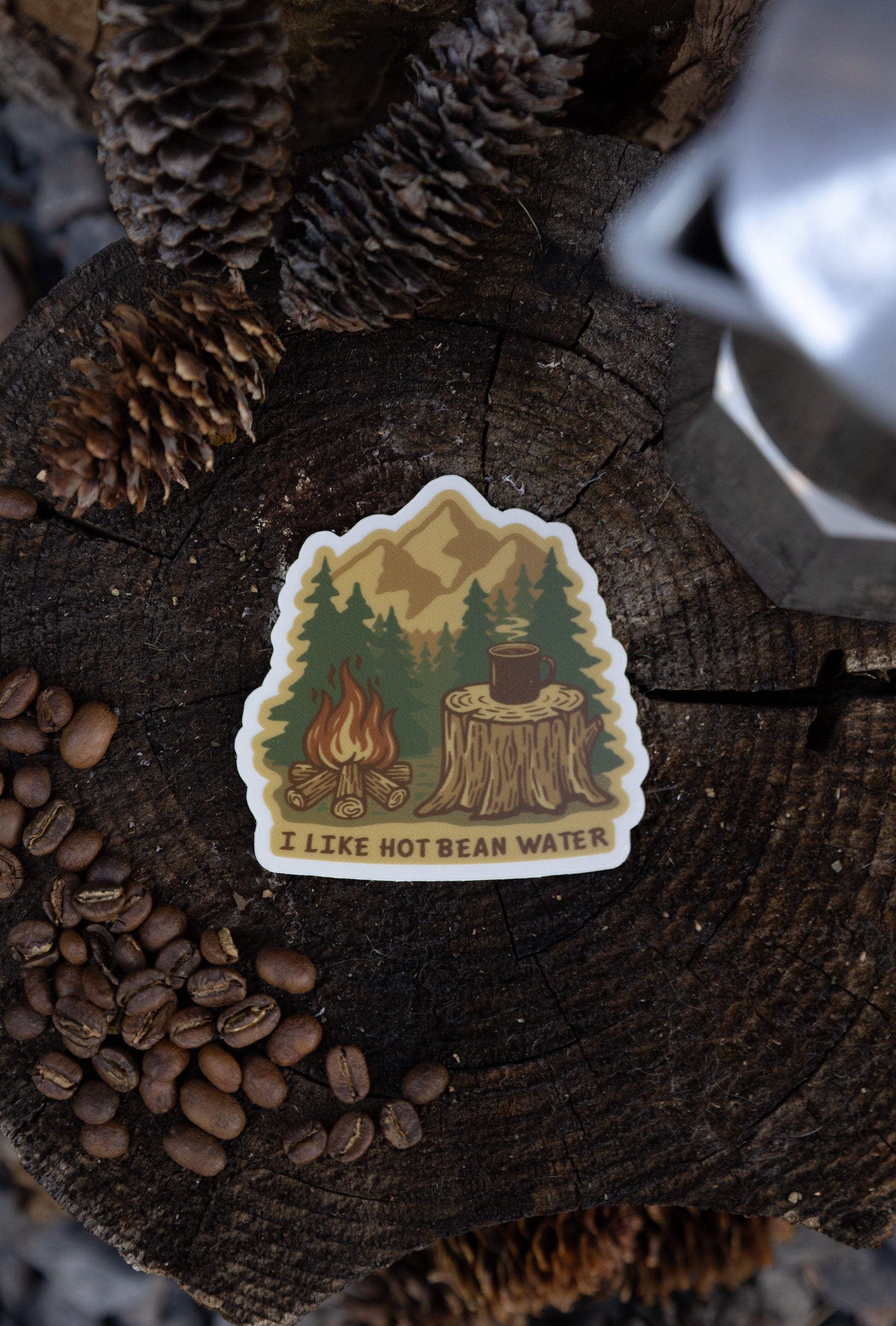 An overhead flat lay of the "I Like Hot Bean Water" sticker resting on a weathered tree stump, surrounded by pinecones, coffee beans, and a silver metal coffee maker.
