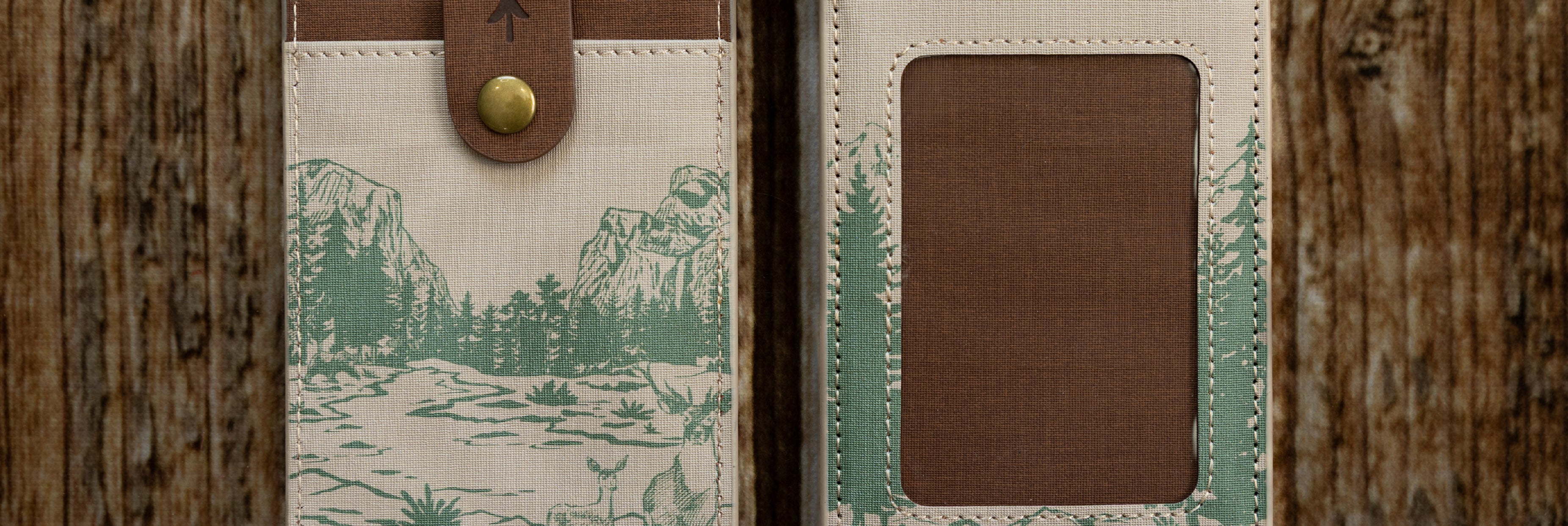 Close-up banner image showcasing the front and back of a cream-colored National Park themed ID holder against a wood background. It highlights detailed green mountain and forest sketches alongside a brown textured window and snap closure.