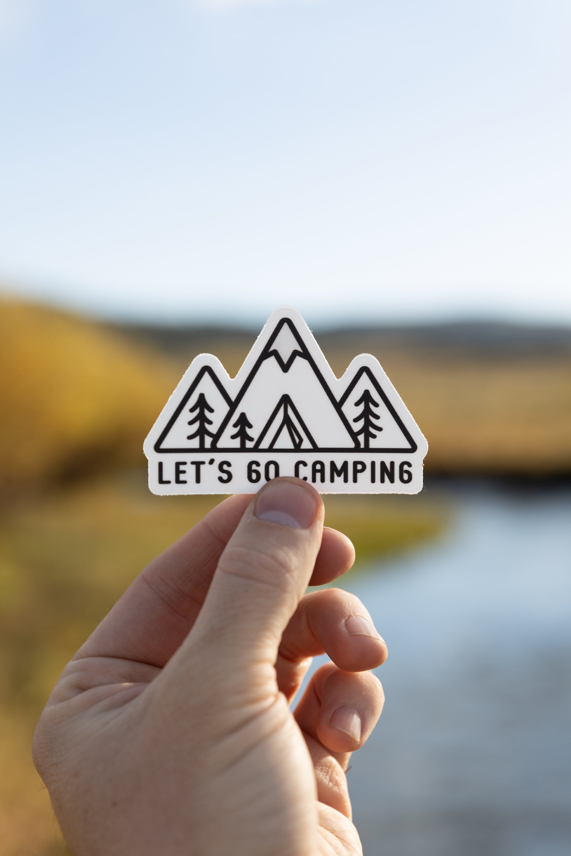 Hand holding out Sticker design of Simplistic line drawing of mountains, tent and trees with the words "Let's go camping" in front of river and valley