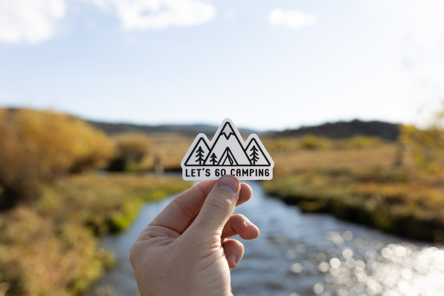 Hand holding out Sticker design of Simplistic line drawing of mountains, tent and trees with the words "Let's go camping" in front of river and valley