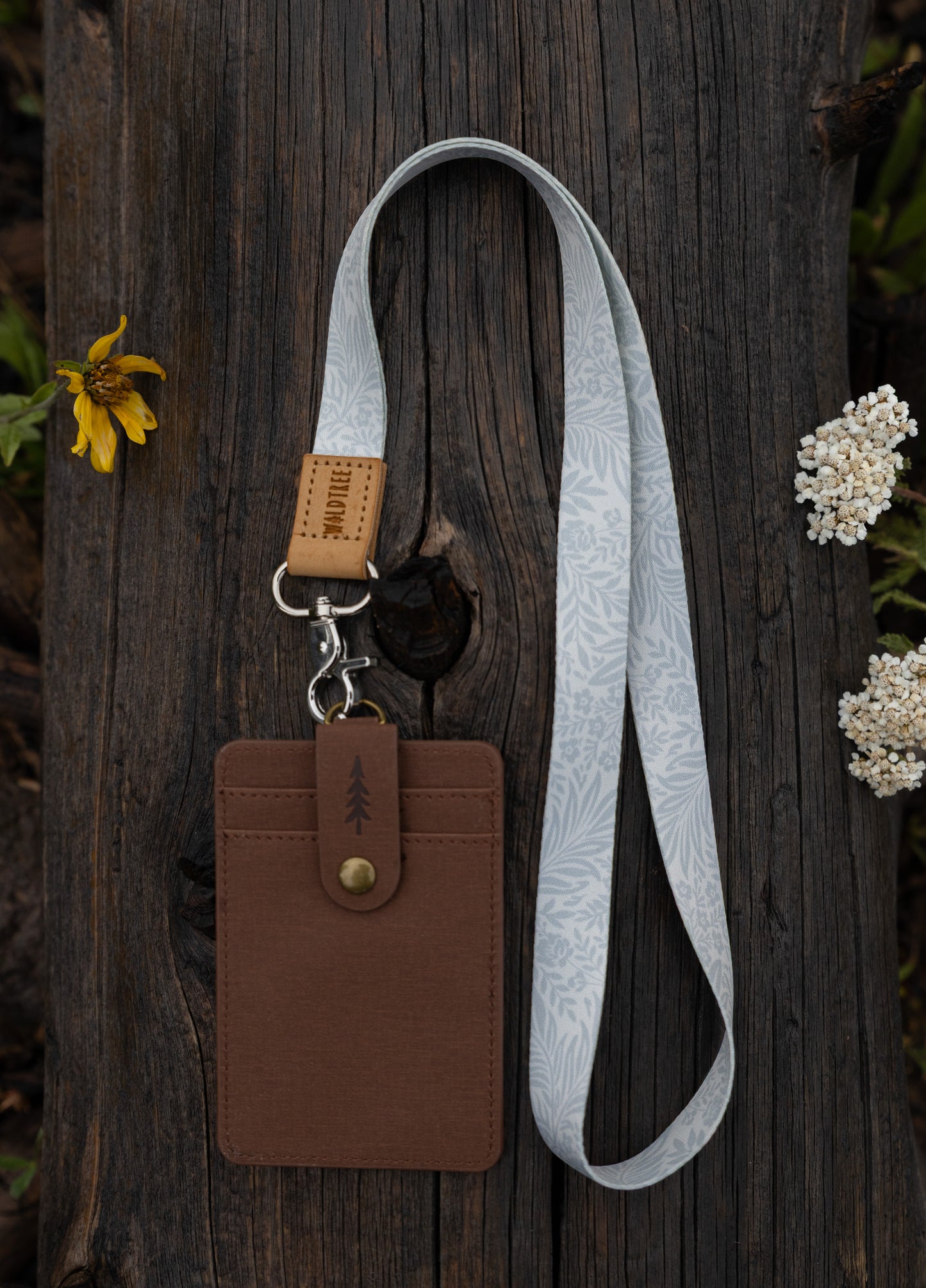 The Meadow Mist neck lanyard laid out on a wooden log surrounded by wildflowers, showing the pale sage fern-print strap, tan leather WILDTREE tab, silver clasp, and a brown vegan-leather card holder attached at the end.