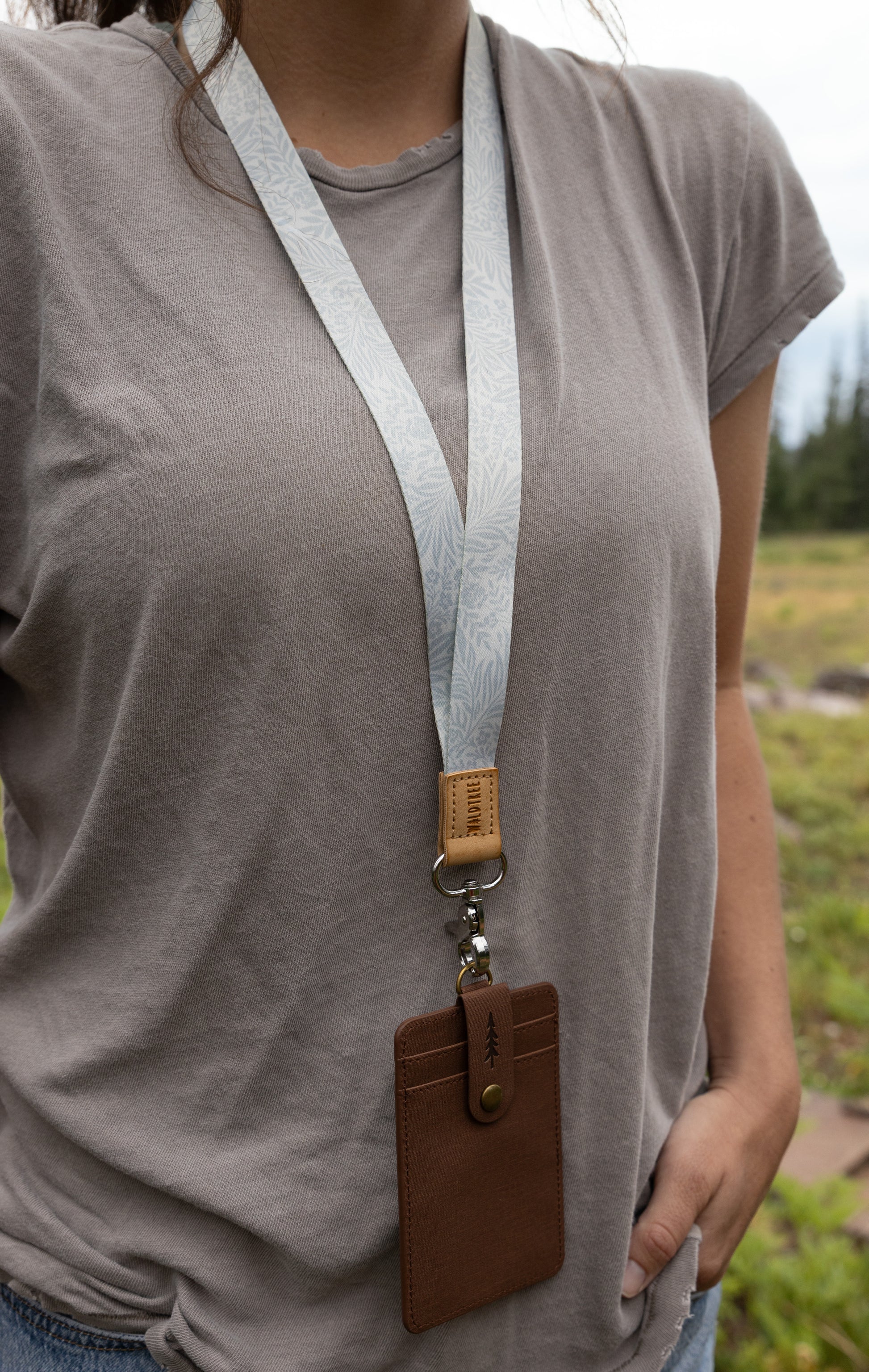 A woman wearing a gray t-shirt models the Meadow Mist neck lanyard, the pale sage fern-print strap leading down to a tan leather tab, silver clasp, and brown vegan-leather card holder, with a meadow and forest in the background.