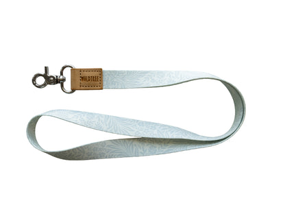 Flat lay of the Meadow Mist neck lanyard on a white background, showing the full pale sage-gray strap printed with a soft white fern leaf pattern, a tan vegan-leather tab embossed with the WILDTREE logo, and a silver swivel clasp.