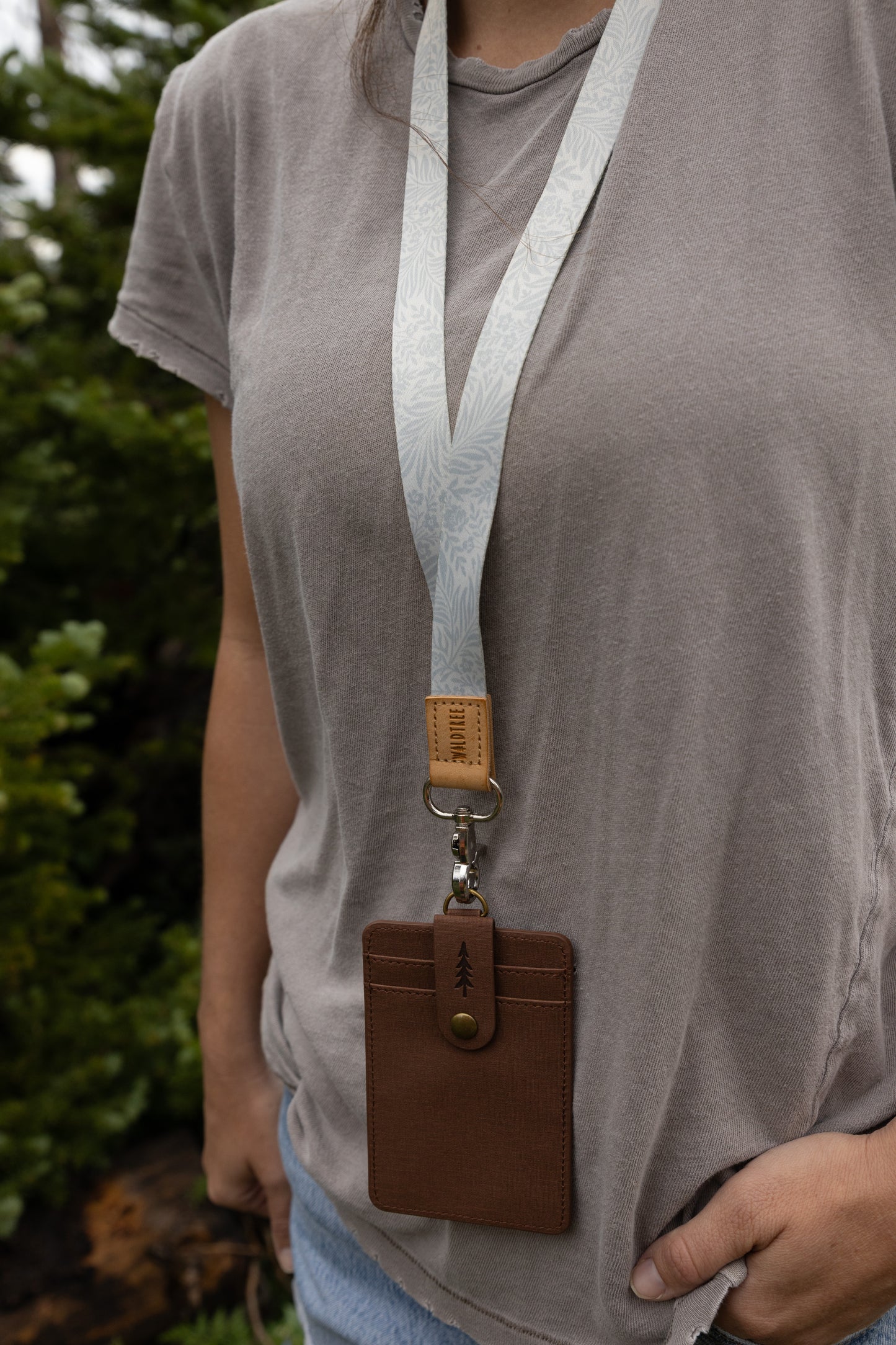 Close-up of the Meadow Mist neck lanyard worn around a woman's neck, showing the pale sage fern-print strap, tan leather WILDTREE tab, silver clasp, and brown vegan-leather card holder against a gray t-shirt.