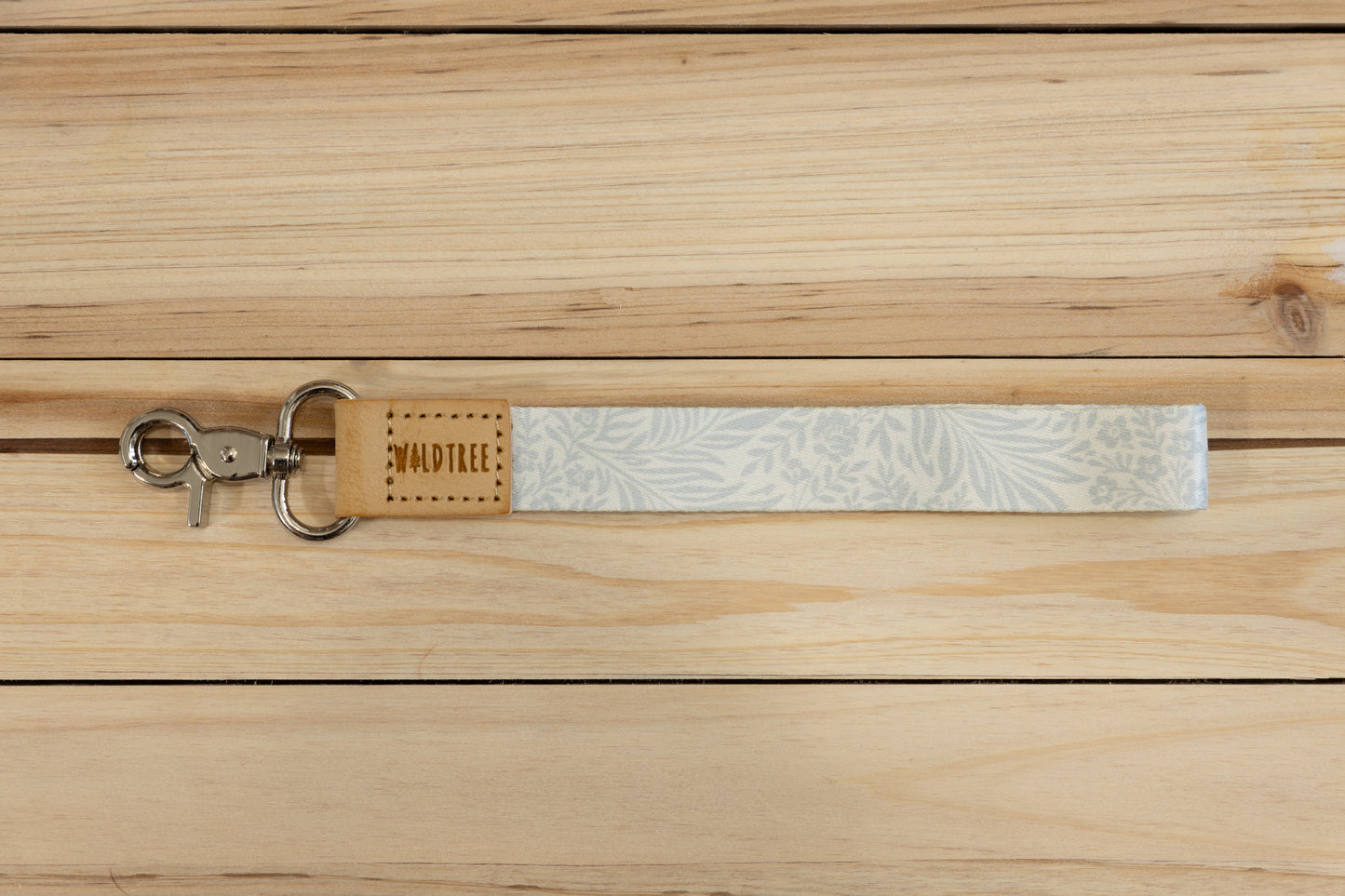 The Meadow Mist wristlet keychain laid flat on a wood plank surface, showing the pale sage fern-print strap, the tan leather WILDTREE tab, and the silver swivel clasp.