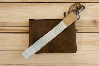 The Meadow Mist wristlet keychain attached to a set of keys and draped across a brown leather zip pouch on a wood surface, showing the pale sage fern-print strap and tan leather WILDTREE tab.