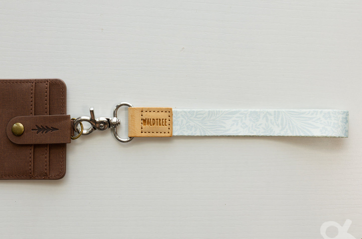 The Meadow Mist wristlet keychain attached to a brown vegan-leather bifold wallet on a white background, showing the pale sage fern-print strap, the tan leather WILDTREE tab, and a silver swivel clasp.