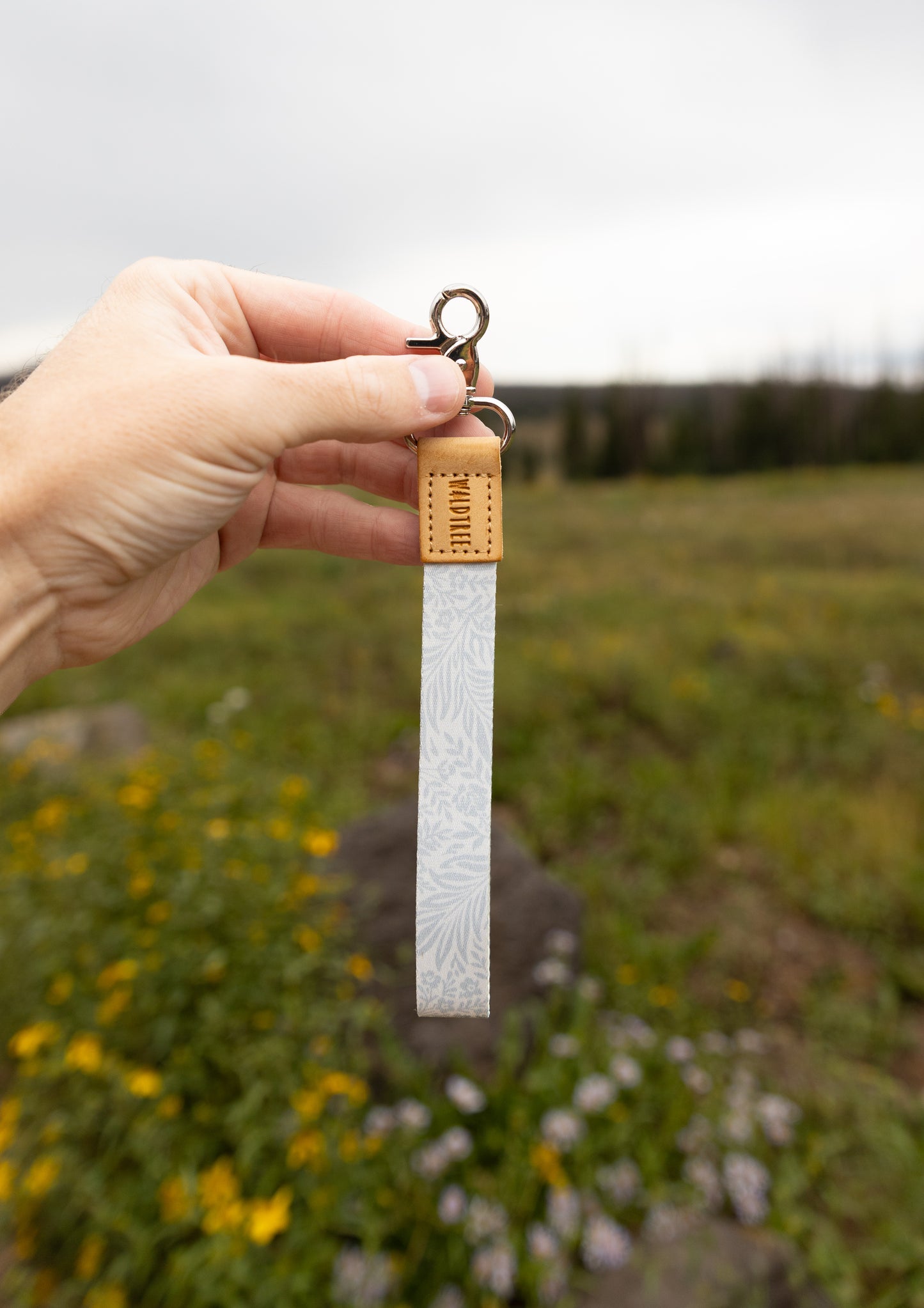 A hand holds the Meadow Mist wristlet keychain up against a cloudy sky and grassy wildflower field, showing the pale sage fern-print strap, the tan leather WILDTREE tab, and silver clasps.