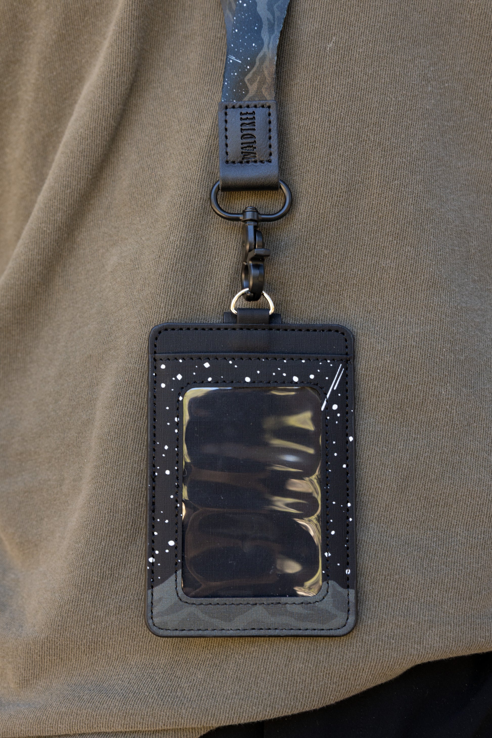 Front and back product layout views of the Midnight Mountain landscape ID badge holder resting flat on a natural wood log outdoors. This descriptive shot clearly highlights the clear front viewing screen on one side and the dual-pocket wallet setup with a brown snap strap on the reverse.